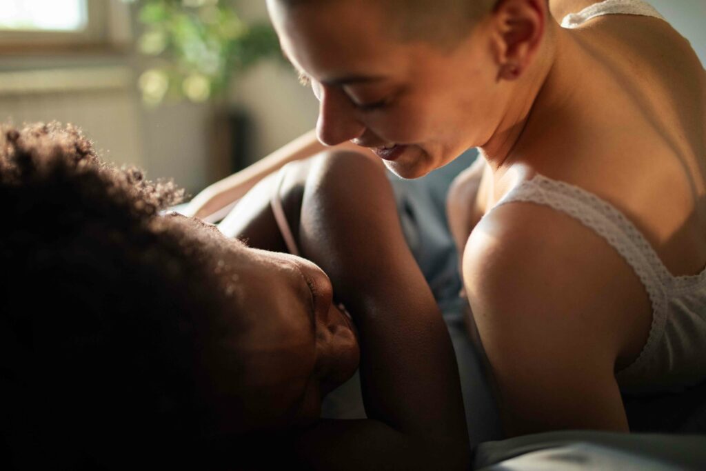 Due donne abbracciate su un letto, in una luce calda; una sorride guardando l’altra