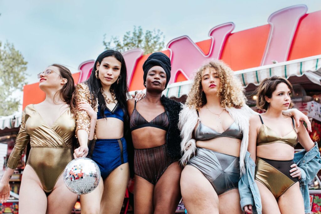 Cinque donne dai corpi diversi, in lingerie metallizzata, posano a braccetto davanti a una giostra del luna park